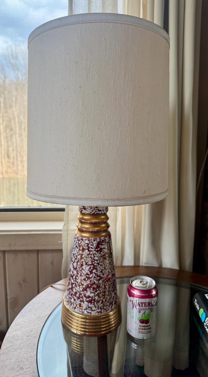May include: A vintage table lamp with a cream-colored lampshade and a decorative base. The base is a deep red color with white and gold accents. The lamp is on a round glass table. A can of Waterloo Cherry Limeade is next to the lamp.