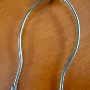 May include: A silver-colored lamp harp with a butterfly-shaped finial. The harp has a U-shaped design with two cylindrical ends. The finial has a screw mechanism for attachment. The lamp harp is designed to hold a lampshade.