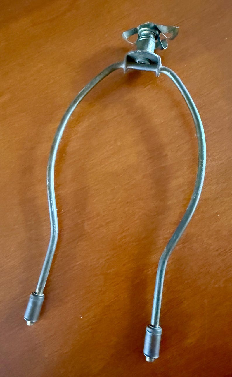 May include: A silver-colored lamp harp with a butterfly-shaped finial. The harp has a U-shaped design with two cylindrical ends. The finial has a screw mechanism for attachment. The lamp harp is designed to hold a lampshade.