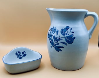 May include: A light gray ceramic pitcher and a matching small bowl. Both pieces feature a hand-painted dark blue floral pattern. The pitcher has a curved handle and a pouring spout, while the bowl has a rounded triangular shape with a blue rim.