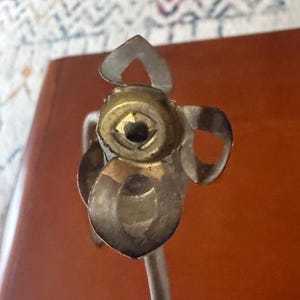 May include: Close-up of a silver and gold-toned metal object with a floral design. The object has a central circular element and petal-shaped details. The stem is visible, suggesting it may be a decorative item or part of a larger piece.