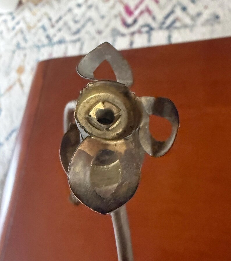 May include: Close-up of a silver and gold-toned metal object with a floral design. The object has a central circular element and petal-shaped details. The stem is visible, suggesting it may be a decorative item or part of a larger piece.