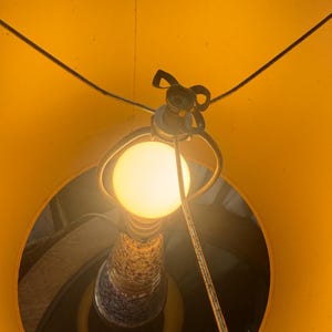May include: A lit lamp viewed from below, showcasing a bright yellow lampshade and a warm, glowing lightbulb. The lamp's base features a speckled design with blue and white accents. The lamp's metal frame is visible.