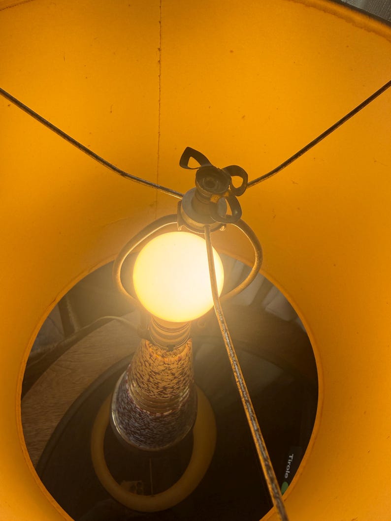 May include: A lit lamp viewed from below, showcasing a bright yellow lampshade and a warm, glowing lightbulb. The lamp's base features a speckled design with blue and white accents. The lamp's metal frame is visible.