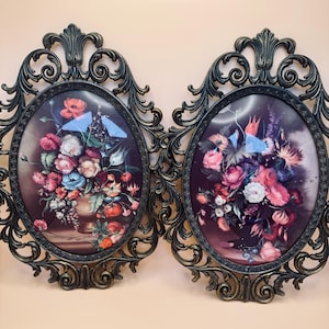 May include: Two ornate, oval-shaped wall art pieces. Each features a floral still life painting in a dark oval frame. The frames are dark bronze with intricate scrollwork. The floral arrangements include red, pink, and blue flowers.