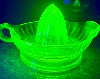 May include: A bright neon green glass citrus juicer featuring a central reamer cone surrounded by a ribbed bowl with a small handle on one side and a pouring spout on the other. The glass glows with a vibrant, translucent fluorescent light.
