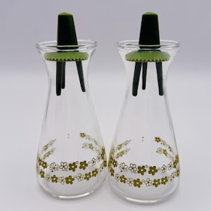 May include: Two clear glass cruet bottles with green floral designs and dark green stoppers. The stoppers have a green base and three black legs. The bottles are designed with a flared base and a narrow neck.