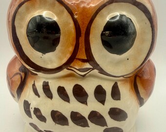 May include: A ceramic owl figurine featuring a cream-colored body with hand-painted brown teardrop-shaped spots. The owl has large, prominent black eyes outlined in dark brown, a small beak, and pointed ear tufts at the top of its head.
