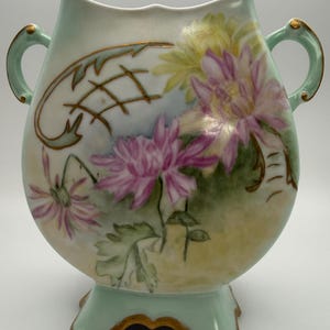 May include: An antique, hand-painted vase with a light green base and handles. The vase features a floral design with pink and yellow flowers, green leaves, and gold accents. The vase has a unique shape and is likely made of porcelain.