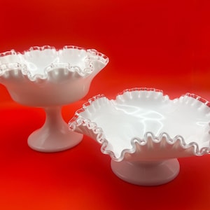 May include: Two white, ruffled-edge glass serving dishes with a red background. One dish is on a pedestal base, the other is a low bowl. The rims have a clear, scalloped edge.