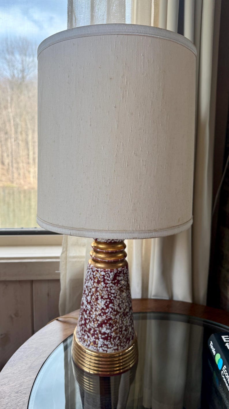 May include: A vintage table lamp with a cream-colored lampshade and a speckled red and white ceramic base. The base has gold-colored accents. The lamp is on a round table near a window.