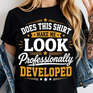 Pode incluir: Uma t-shirt preta de manga curta com texto em branco e amarelo que diz: DOES THIS SHIRT MAKE ME LOOK Professionally DEVELOPED. O design inclui pequenos detalhes de estrelas e linhas horizontais, centrado na frente da peça.