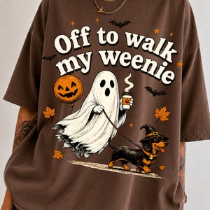 Puede incluir: Una camiseta marrón con un gráfico de un fantasma que sostiene un globo de calabaza y una taza de café, paseando a un perro salchicha con sombrero de bruja. El texto "Off to walk my weenie" está rodeado de hojas de arce naranjas, estrellas y murciélagos.