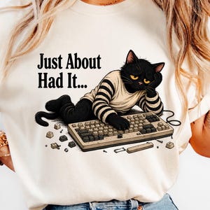 May include: A cream-colored t-shirt featuring a graphic of a black cat wearing a striped shirt, resting its head on a hand while typing on a computer keyboard. The text "Just About Had It..." is printed above the cat, with scattered keyboard keys.