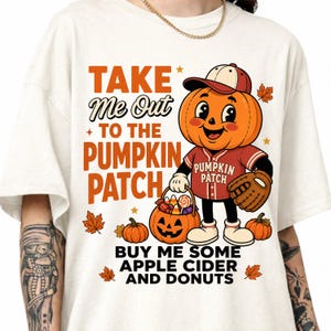 May include: An off-white t-shirt featuring a graphic of a cartoon pumpkin character wearing a baseball cap and jersey, holding a candy bucket. The text reads: TAKE ME OUT TO THE PUMPKIN PATCH, BUY ME SOME APPLE CIDER AND DONUTS, with autumn leaves.