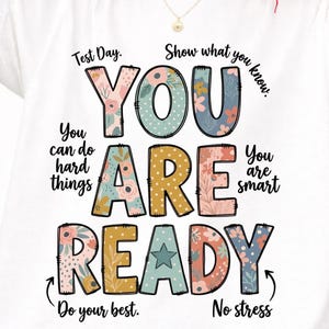Può includere: T-shirt bianca con le parole "YOU ARE READY" in grandi lettere colorate con motivi floreali. La maglietta include anche le frasi "Test Day", "Show what you know", "You can do hard things", "You are smart", "Do your best" e "No stress".