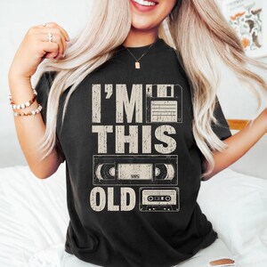 May include: Black t-shirt with the text "I'M THIS OLD" above a floppy disk, VHS tape, and cassette tape graphic. The design has a distressed, vintage look.