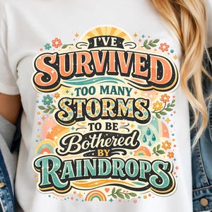 以下が含まれることがあります： カラフルなグラフィックデザインの白いTシャツ。デザインは、レトロなフォントで「I've survived too many storms to be bothered by raindrops」というテキストを特徴とし、花柄と虹色のアクセントが付いています。シャツは柔らかい素材で作られています。