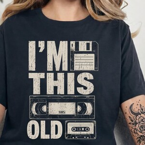 May include: Black t-shirt with the text "I'M THIS OLD" in distressed white lettering. The design includes illustrations of a floppy disk, a VHS tape, and a cassette tape, evoking a retro theme.
