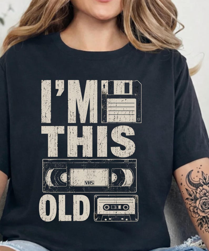 May include: Black t-shirt with the text "I'M THIS OLD" in distressed white lettering. The design includes illustrations of a floppy disk, a VHS tape, and a cassette tape, evoking a retro theme.