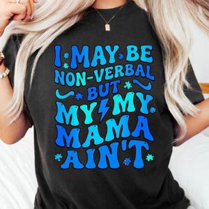 Puede incluir: Camiseta negra con el texto "I MAY BE NON-VERBAL BUT MY MAMA AIN'T" en letras azules y celeste. La camiseta tiene cuello redondo y mangas cortas. El texto incluye gráficos de piezas de rompecabezas.