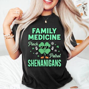 以下が含まれることがあります： 黒いTシャツ。緑色の文字で「FAMILY MEDICINE」と書かれています。サングラスとオレンジ色の靴を履いた緑色のクローバーが中央にあります。「Pinch Patrol Shenanigans」の文字もプリントされています。