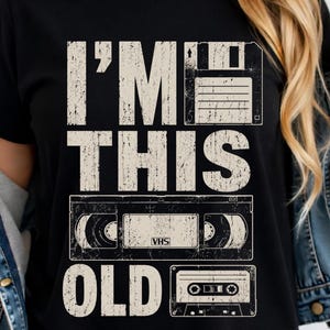 May include: Black t-shirt with a distressed white graphic. The graphic features the text "I'M THIS OLD" with illustrations of a floppy disk, a VHS tape, and a cassette tape, evoking a retro theme.