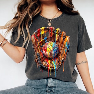 May include: Dark gray t-shirt featuring a baseball glove and ball with colorful paint drips. The baseball is covered in sprinkles. The glove is brown with black stitching. The shirt is a crew neck.