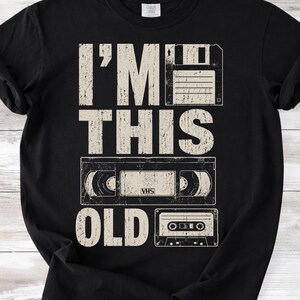 May include: Black t-shirt with the text "I'M THIS OLD" in a distressed, off-white font. The design features a floppy disk, a VHS tape, and a cassette tape, evoking a retro theme.