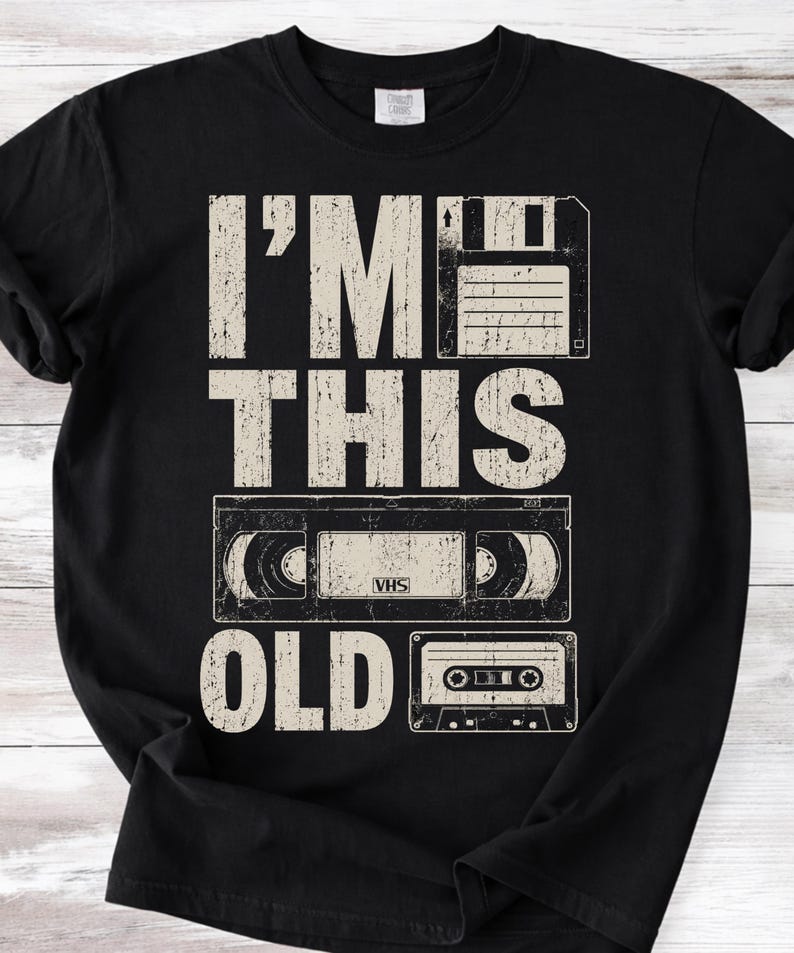 May include: Black t-shirt with the text "I'M THIS OLD" in a distressed, off-white font. The design features a floppy disk, a VHS tape, and a cassette tape, evoking a retro theme.