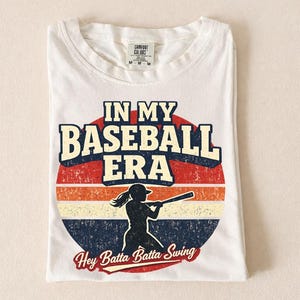 May include: White t-shirt with the text "IN MY BASEBALL ERA" in a retro design. The design includes a silhouette of a batter and the phrase "Hey Batta Batta Swing". The shirt is folded and has a tag that says "Comfort Colors".