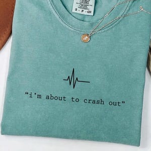以下が含まれることがあります： 「i'm about to crash out」のテキストと黒い心拍ラインのグラフィックがプリントされた、ミントグリーンのTシャツ。小さなペンダントが付いたシルバーのネックレスがシャツの上に置かれています。