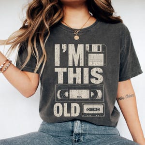 May include: Dark gray t-shirt with the text "I'M THIS OLD" above a floppy disk, VHS tape, and cassette tape graphic. The shirt has a vintage, worn look.