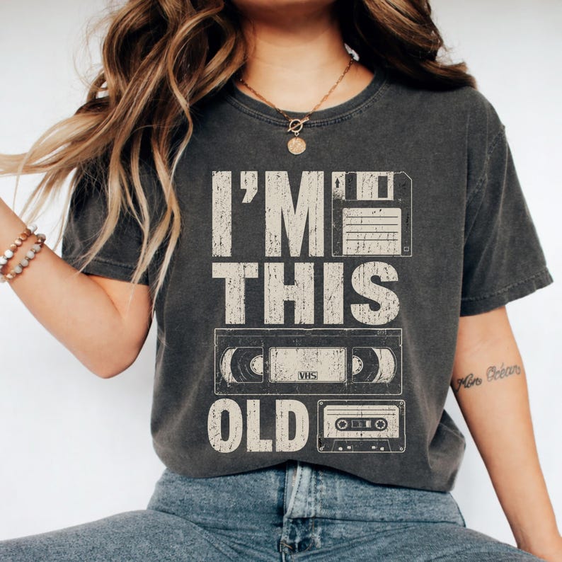 May include: Dark gray t-shirt with the text "I'M THIS OLD" above a floppy disk, VHS tape, and cassette tape graphic. The shirt has a vintage, worn look.