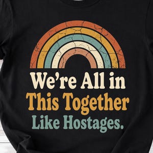 Puede incluir: Camiseta negra con un diseño de arcoíris retro en tonos naranja, amarillo, azul y crema. El texto de la camiseta dice "We're All in This Together Like Hostages" en una fuente vintage.