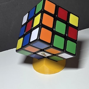 May include: A Rubik's Cube with black edges and colorful squares in red, orange, yellow, green, blue, and white. The cube is resting on a yellow stand. The cube is a classic puzzle toy.