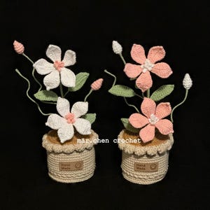 Könnte beinhalten: Zwei gehäkelte Blumenarrangements. Eines zeigt weiße Blüten mit rosa Zentren, das andere rosa Blüten mit weißen Zentren. Beide befinden sich in beigefarbenen Töpfen mit "Hand Made"-Etiketten und dem Text "mär chen crochet".