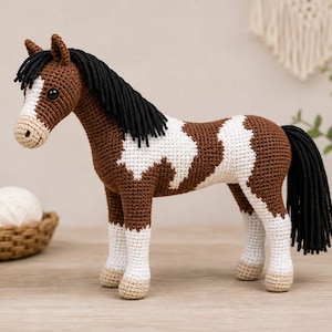 May include: A brown and white crocheted horse toy with a black mane and tail. The horse features a white blaze on its face, white patches on its body, and white lower legs with light tan hooves. The toy has dark eyes and a stitched mouth.