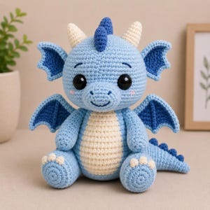May include: A light blue crocheted dragon with dark blue wings, a matching fin, and cream-colored horns. The dragon has large black eyes, a cream-colored belly, and small pink cheeks. The dragon is sitting down.