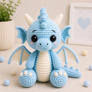 May include: A light blue crocheted dragon with white accents. The handmade plush toy features large black eyes, small pink cheeks, and white claws. The dragon has wings and is sitting on a surface with small blue and white balls.