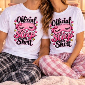 Könnte beinhalten: Zwei weiße kurzärmelige T-Shirts mit einem Grafikdruck, der Official Sleep Shirt in schwarzer und rosa Schrift zeigt. Das Design enthält eine rosa Schlafmaske mit einer Schleife, umgeben von kleinen rosa Herzen und gelben Sternen.