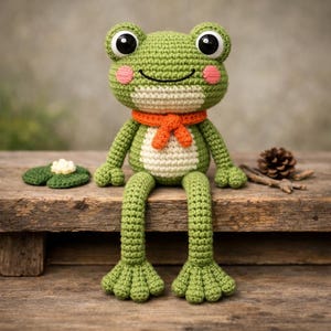 Op de afbeelding: Een handgemaakte groene gehaakte kikkerpop met grote zwarte ogen, roze wangen en een oranje sjaal. De kikker zit met zijn lange benen te bungelen. Een gehaakte waterlelie en een dennenappel liggen in de buurt.