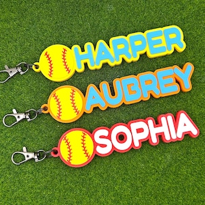 May include: Three custom keychains featuring a yellow softball icon with red stitching attached to a metal lobster clasp. Each keychain displays a name in raised block letters: HARPER in blue, AUBREY in blue, and SOPHIA in white with a red border.