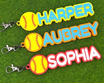 Op de afbeelding: Drie gepersonaliseerde sleutelhangers met een geel softbal-icoon met rode stiksels, bevestigd aan een metalen karabijnhaak. Elke sleutelhanger toont een naam in verhoogde blokletters: HARPER in blauw, AUBREY in blauw en SOPHIA in wit met een rode rand.
