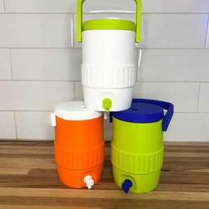 May include: Three colorful water coolers stacked on a wooden surface. The coolers are white, orange, and green, each with a matching lid and handle. The white cooler has a green handle, the orange has a white lid, and the green has a blue lid.
