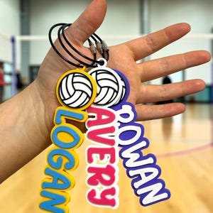 May include: Three custom volleyball keychains attached to black metal rings. Each keychain features a white volleyball icon at the top and a name in raised lettering: Logan in blue on yellow, Avery in pink on white, and Rowan in purple on white.