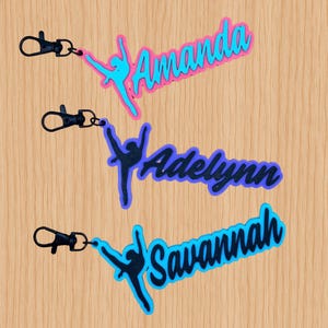 May include: Three custom keychains featuring a silhouette of a dancer in a split pose attached to a black lobster clasp. The top keychain is pink and blue with the name Amanda, the middle is purple with Adelynn, and the bottom is blue with Savannah.