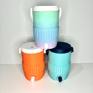 May include: Three colorful water coolers stacked on a white surface. The top cooler has a gradient of green to blue, the middle one is orange, and the bottom one is teal. Each cooler has a white handle and a spigot.