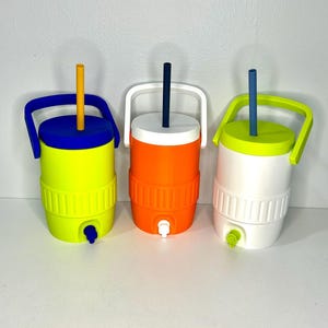 May include: Three colorful water coolers with lids and handles. The coolers are yellow with a blue lid and handle, orange with a white lid and handle, and white with a green lid and handle. Each has a straw and a spigot.