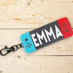 May include: A rectangular keychain shaped like a game controller with a black center, a light blue left side, and a red right side. The name EMMA is printed in large white letters across the black center. A black metal lobster clasp is attached.